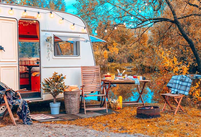 Vintage camper trailer campsite beside a calm lake in an autumn forest, with string lights, outdoor chairs, and a picnic table. Vintage camper trailer campsite beside a calm lake in an autumn forest, with string lights, outdoor chairs, and a picnic table.